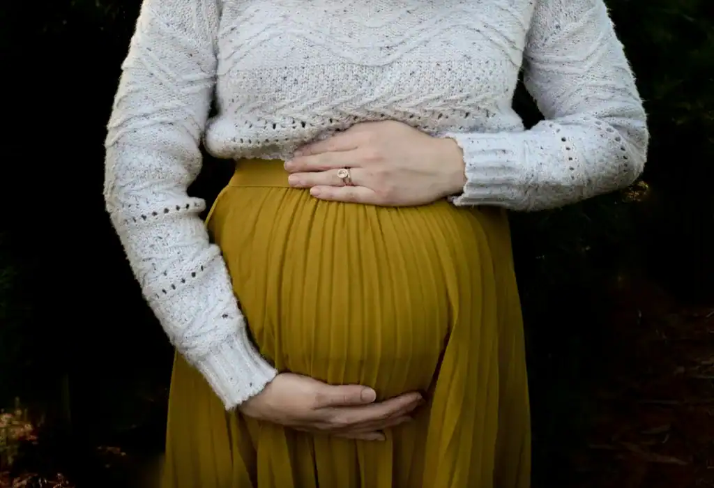 How To Glow-Up Postpartum: 5 Super Realistic Ways to Feel Like Yourself Again 9 faceless woman in a white shirt and yellow skirt holding her pregnancy bump.